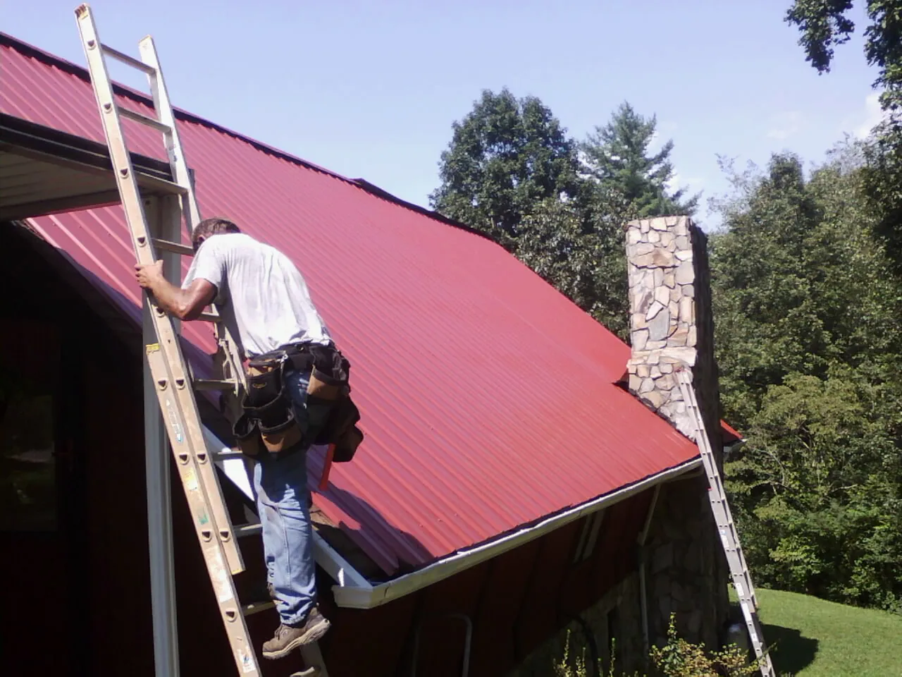 Roofer on a ladder beside a red metal roof and stone chimney — Soffit & Fascia Repair by Roofing Brothers in Oil City